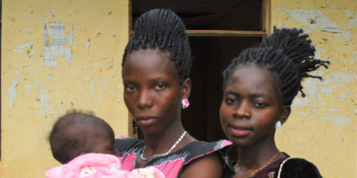 Bénéficiaire JRS d’une bourse de formation à l’enseignement, Evelyn porte la nouvelle-née de Nakio tandis qu’elles posent pour une photo devant leur salle de classe de l’université Mikese à Yambio, Soudan du Sud. (Service Jésuite des Réfugiés)
