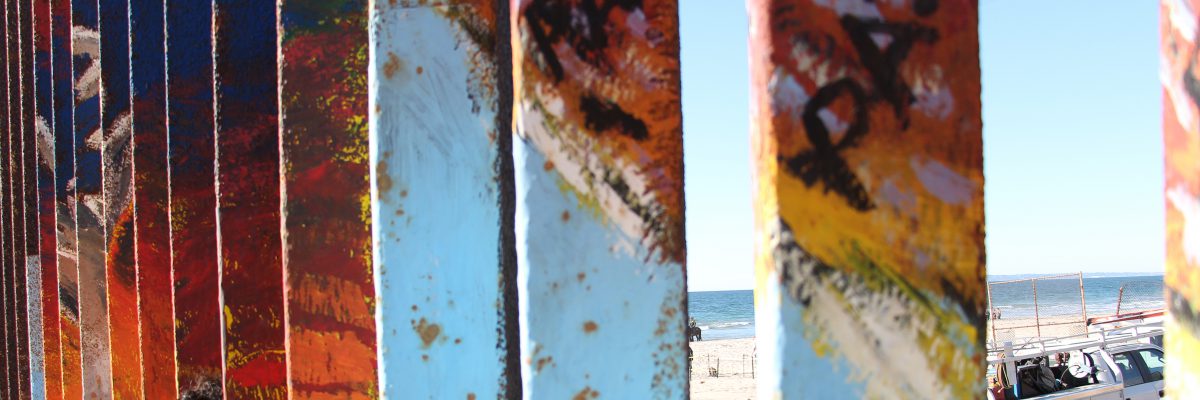 Migrants at the border between Tijuana and San Diego. The border fence is painted with the word