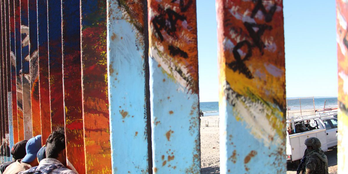 Migrants at the border between Tijuana and San Diego. The border fence is painted with the word 