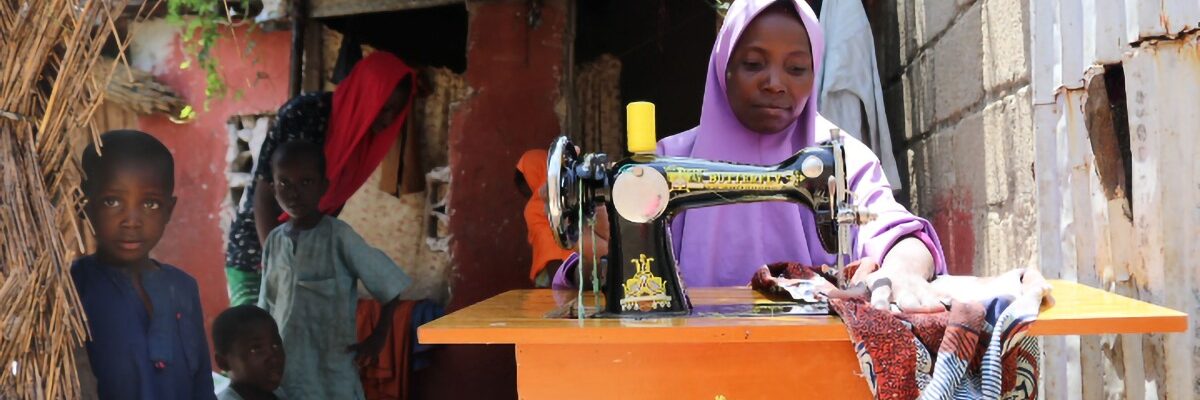 Grâce au financement humanitaire de l'Union Européenne (UE), le JRS fournit une aide aux moyens de subsistance aux femmes nigérianes. Fátima coud des vêtements pour un client chez elle, entourée de ses enfants. État de Borno, Nigeria (Service jésuite des réfugiés).