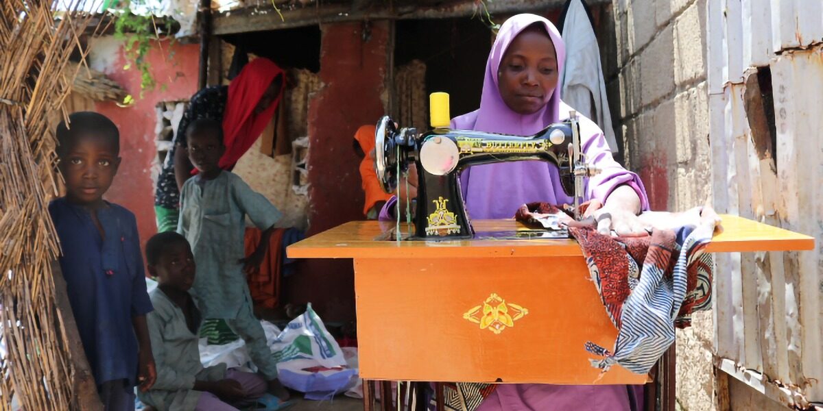 Grâce au financement humanitaire de l'Union Européenne (UE), le JRS fournit une aide aux moyens de subsistance aux femmes nigérianes. Fátima coud des vêtements pour un client chez elle, entourée de ses enfants. État de Borno, Nigeria (Service jésuite des réfugiés).